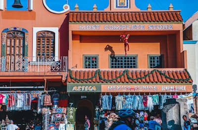 A vibrant street scene features a colorful building with terracotta roofing. The facade has decorative windows and signage reading 'Most Complete Store' and 'Mexico Curios.' An Iron Man figurine is attached to an upper balcony. Below, clothing and souvenirs are displayed outside the store, with people browsing.