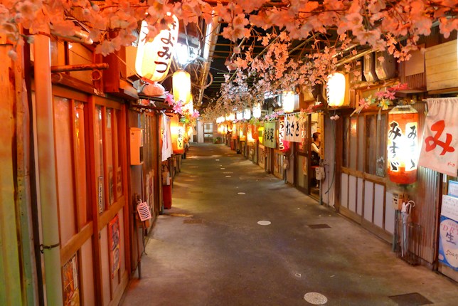 A narrow alley in a traditional Japanese street lined with wooden structures and vibrant paper lanterns. Cherry blossom decorations hang overhead, creating a festive atmosphere. The warm glow from the lanterns and shop signs illuminates the path.