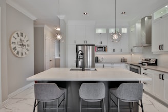 A modern kitchen with white cabinets, stainless steel appliances, and a central island with a sink. Three grey bar stools are positioned at the island, which is illuminated by two geometric pendant lights. A round clock with Roman numerals decorates the wall, and the overall decor conveys a sleek and contemporary design.