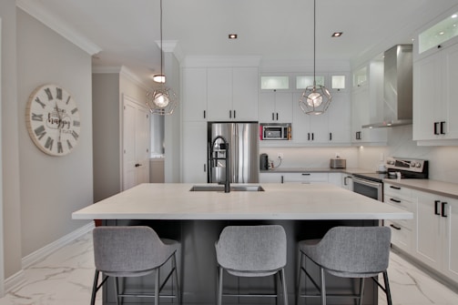 A modern kitchen with white cabinets, stainless steel appliances, and a central island with a sink. Three grey bar stools are positioned at the island, which is illuminated by two geometric pendant lights. A round clock with Roman numerals decorates the wall, and the overall decor conveys a sleek and contemporary design.