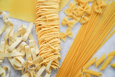 Various types of uncooked pasta are neatly arranged in an aesthetically pleasing manner. The assortment includes fettuccine, spaghetti, bow-tie, lasagna sheets, and penne. The pasta is set against a light textured surface, highlighting its golden-yellow hue.