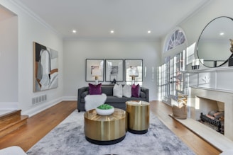 A stylish, contemporary living room featuring a comfortable gray sofa with decorative cushions and a soft throw. Two modern gold-toned coffee tables are set on a plush gray rug. The space is accented by wall art, a large mirror above the fireplace, and elegant table lamps. Natural light filters through tall glass doors, highlighting the detailed decor.