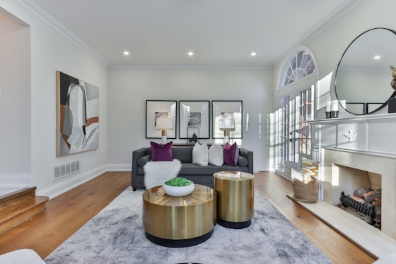 A stylish, contemporary living room featuring a comfortable gray sofa with decorative cushions and a soft throw. Two modern gold-toned coffee tables are set on a plush gray rug. The space is accented by wall art, a large mirror above the fireplace, and elegant table lamps. Natural light filters through tall glass doors, highlighting the detailed decor.