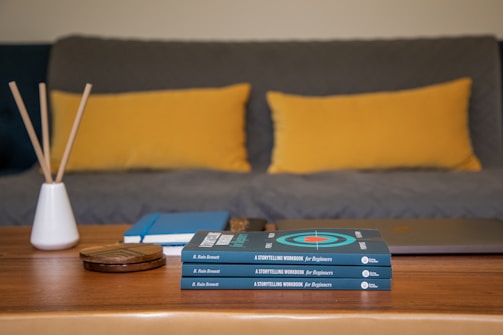 A wooden coffee table is topped with a pile of books titled 'A Storytelling Workbook for Beginners' by B. Rain Bennett. Beside the books is a closed laptop, a few notebooks, and a small white vase holding incense sticks. In the background, a gray couch is adorned with two large mustard-yellow pillows.