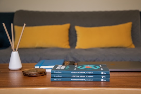 A wooden coffee table is topped with a pile of books titled 'A Storytelling Workbook for Beginners' by B. Rain Bennett. Beside the books is a closed laptop, a few notebooks, and a small white vase holding incense sticks. In the background, a gray couch is adorned with two large mustard-yellow pillows.