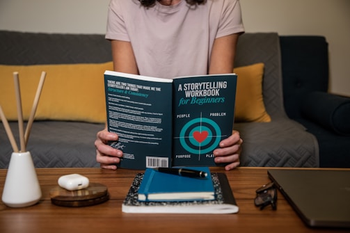A person holding a book titled 'A Storytelling Workbook for Beginners' while sitting on a sofa. The book cover features a heart design with the words 'people,' 'problem,' 'purpose,' and 'plan.' The setting includes a coffee table with a notebook, pen, and eyeglasses, and a decorative diffuser in the foreground. The sofa has a gray quilted texture and a yellow pillow.