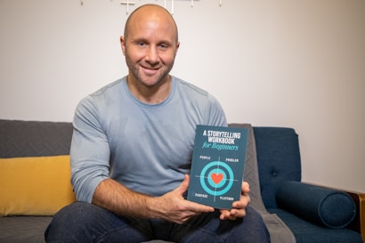 A man is sitting on a couch holding a book titled 'A Storytelling Workbook for Beginners.' The book cover features a design with concentric circles and the words 'People,' 'Problem,' 'Purpose,' and 'Platform' with a heart at the center. The man is wearing a light blue sweater and smiling at the camera. The couch has a yellow cushion and is in a room with a minimalist style.