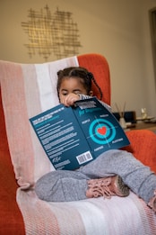 A young child with braided hair sits on a soft, orange armchair, covered with a light, stripy blanket. The child is holding a book titled 'A Storytelling Workbook for Beginners' with a blue cover featuring a heart inside a target graphic. The focus is on the child and the book, with some modern decor visible in the background.
