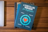 A blue book titled 'A Storytelling Workbook for Beginners' is placed on top of a composition-style notebook with a marbled black and white cover. The book features a target graphic with a red heart in the center and the words surrounding it are 'People', 'Problem', 'Purpose', and 'Platform'. Beneath this stack, the corner of a silver laptop is visible on a wooden surface.