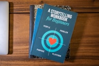 A blue book titled 'A Storytelling Workbook for Beginners' is placed on top of a composition-style notebook with a marbled black and white cover. The book features a target graphic with a red heart in the center and the words surrounding it are 'People', 'Problem', 'Purpose', and 'Platform'. Beneath this stack, the corner of a silver laptop is visible on a wooden surface.