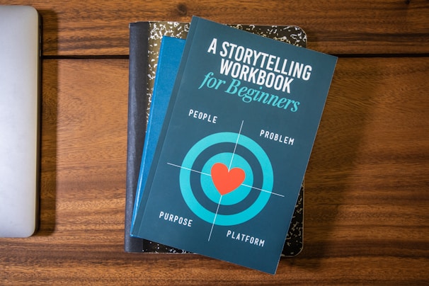 A blue book titled 'A Storytelling Workbook for Beginners' is placed on top of a composition-style notebook with a marbled black and white cover. The book features a target graphic with a red heart in the center and the words surrounding it are 'People', 'Problem', 'Purpose', and 'Platform'. Beneath this stack, the corner of a silver laptop is visible on a wooden surface.