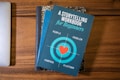 A blue book titled 'A Storytelling Workbook for Beginners' is placed on top of a composition-style notebook with a marbled black and white cover. The book features a target graphic with a red heart in the center and the words surrounding it are 'People', 'Problem', 'Purpose', and 'Platform'. Beneath this stack, the corner of a silver laptop is visible on a wooden surface.