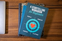 A blue book titled 'A Storytelling Workbook for Beginners' is placed on top of a composition-style notebook with a marbled black and white cover. The book features a target graphic with a red heart in the center and the words surrounding it are 'People', 'Problem', 'Purpose', and 'Platform'. Beneath this stack, the corner of a silver laptop is visible on a wooden surface.