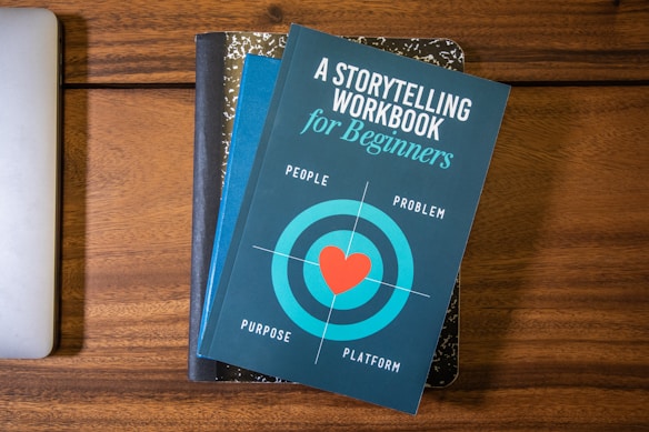 A blue book titled 'A Storytelling Workbook for Beginners' is placed on top of a composition-style notebook with a marbled black and white cover. The book features a target graphic with a red heart in the center and the words surrounding it are 'People', 'Problem', 'Purpose', and 'Platform'. Beneath this stack, the corner of a silver laptop is visible on a wooden surface.