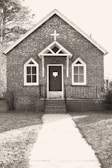 A small, rustic brick church with Gothic-style arched windows and a prominent cross above the entrance. The path leading to the front door is lined with grass on either side, and trees are visible in the background.