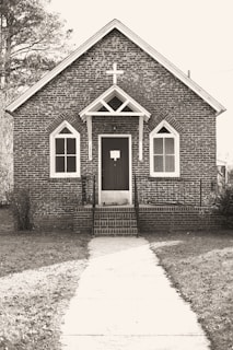 A small, rustic brick church with Gothic-style arched windows and a prominent cross above the entrance. The path leading to the front door is lined with grass on either side, and trees are visible in the background.