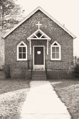 A small, rustic brick church with Gothic-style arched windows and a prominent cross above the entrance. The path leading to the front door is lined with grass on either side, and trees are visible in the background.