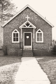 A small, rustic brick church with Gothic-style arched windows and a prominent cross above the entrance. The path leading to the front door is lined with grass on either side, and trees are visible in the background.