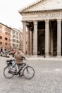 A person stands with a bicycle in front of an ancient Roman building with large columns. The structure has an inscription on its facade and is surrounded by a cobblestone plaza. Several people are seen in the background walking or seated.