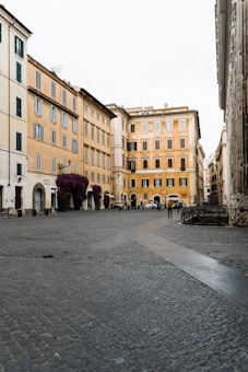 A quaint Italian street scene with historic yellow and light-colored buildings lining a cobbled plaza. A few pedestrians and parked cars are visible, contributing to the serene and nostalgic atmosphere. Bougainvillea flowers adorn the facades, adding a touch of vibrant color.