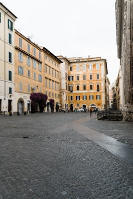 A quaint Italian street scene with historic yellow and light-colored buildings lining a cobbled plaza. A few pedestrians and parked cars are visible, contributing to the serene and nostalgic atmosphere. Bougainvillea flowers adorn the facades, adding a touch of vibrant color.