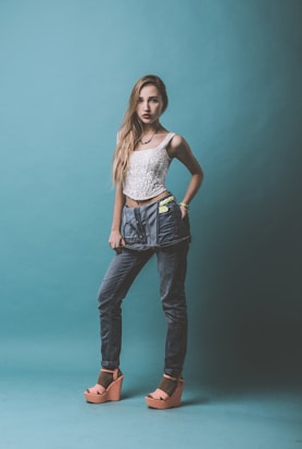 A young woman stands confidently against a teal background. She is wearing a white lace top, high-waisted blue jeans with a unique belt, and peach platform sandals. Her hair is long and straight, falling over one shoulder, and she has a serious expression.