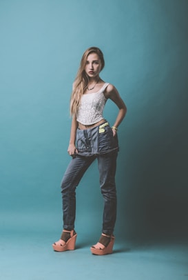 A young woman stands confidently against a teal background. She is wearing a white lace top, high-waisted blue jeans with a unique belt, and peach platform sandals. Her hair is long and straight, falling over one shoulder, and she has a serious expression.