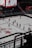 A group of hockey players in red uniforms are practicing or preparing for a game on an indoor ice rink. The arena is mostly empty, with a few spectators visible in the stands. A large digital screen displays images of players on the ice. The ice surface is marked with team logos and advertisements.