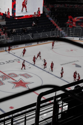 A group of hockey players in red uniforms are practicing or preparing for a game on an indoor ice rink. The arena is mostly empty, with a few spectators visible in the stands. A large digital screen displays images of players on the ice. The ice surface is marked with team logos and advertisements.