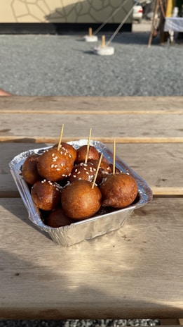 A foil container of golden-brown fried balls, topped with sesame seeds, placed on a wooden picnic table. Toothpicks are inserted in some of the balls. The background includes a gravel area and a modern building with a geometric pattern.