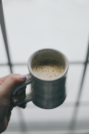 A hand holding a patterned mug filled with steaming coffee by a sunny window.