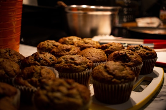 A close-up view of several freshly baked muffins placed on a white tray. The muffins have a golden brown color with visible chunks of chocolate or nuts. In the background, there's a blurred metallic mixing bowl and some kitchen utensils, suggesting a domestic kitchen setting. Warm lighting enhances the cozy and inviting atmosphere.