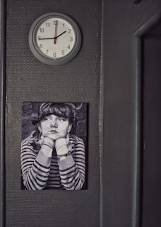 A wall-mounted clock shows the time as 12:05, and below it there is a black and white photo of a person with their hands resting under their chin. The person is wearing a striped shirt and has a contemplative expression.