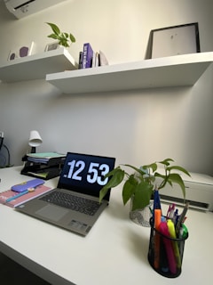 A modern and tidy workspace featuring a white desk with a laptop displaying the time 12:53. On the desk, there is a plant in a decorative pot, a pen holder containing colorful pens, and a neat stack of stationery. Two white shelves are mounted on the wall, holding books, a framed picture, and decorations. The room has a minimalist design with neutral colors.