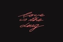 Red neon script handwriting displays the phrase 'love is the drug' against a black background.