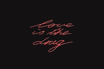 Red neon script handwriting displays the phrase 'love is the drug' against a black background.