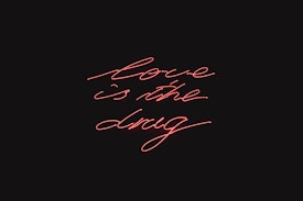 Red neon script handwriting displays the phrase 'love is the drug' against a black background.
