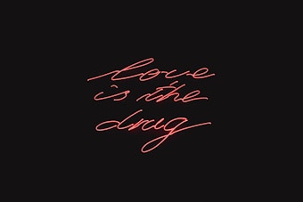 Red neon script handwriting displays the phrase 'love is the drug' against a black background.