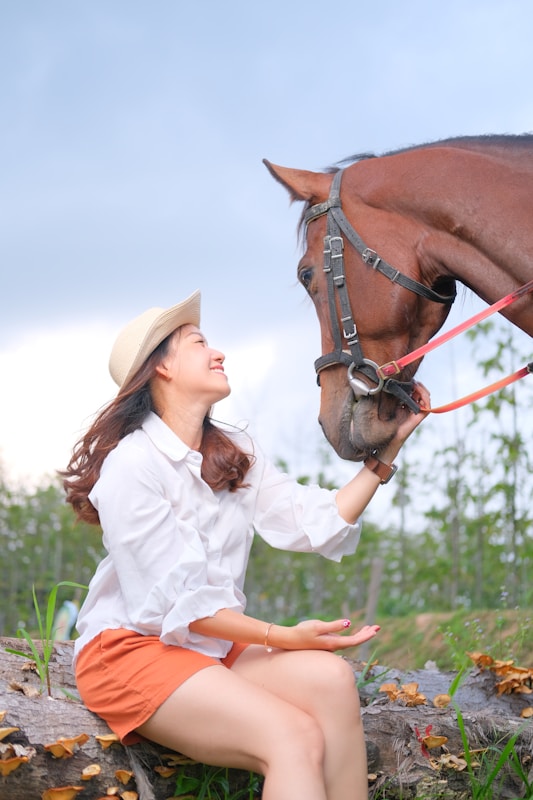A woman wearing an off-white hat, a white shirt, and orange shorts is sitting on a tree log and smiling warmly as she gently pats the head of a brown horse. The background is a natural setting with greenery and a cloudy sky.