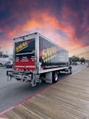 A delivery truck featuring 'SAVAL FOODSERVICE' branding is parked on a wooden boardwalk. The sky displays a vibrant, colorful sunset with shades of orange, pink, and purple. Nearby, there are a few parked cars and trees in the background.