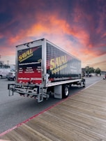 A delivery truck featuring 'SAVAL FOODSERVICE' branding is parked on a wooden boardwalk. The sky displays a vibrant, colorful sunset with shades of orange, pink, and purple. Nearby, there are a few parked cars and trees in the background.