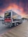 A delivery truck featuring 'SAVAL FOODSERVICE' branding is parked on a wooden boardwalk. The sky displays a vibrant, colorful sunset with shades of orange, pink, and purple. Nearby, there are a few parked cars and trees in the background.