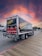 A delivery truck featuring 'SAVAL FOODSERVICE' branding is parked on a wooden boardwalk. The sky displays a vibrant, colorful sunset with shades of orange, pink, and purple. Nearby, there are a few parked cars and trees in the background.
