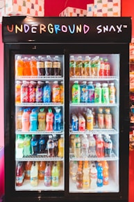A display refrigerator filled with various bottled beverages, including sodas and juices, with a neon 'Underground Snax' sign on top. The bottles are organized neatly on several shelves, each containing different brands and colors.
