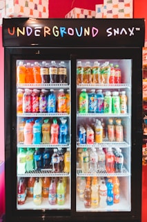 A display refrigerator filled with various bottled beverages, including sodas and juices, with a neon 'Underground Snax' sign on top. The bottles are organized neatly on several shelves, each containing different brands and colors.