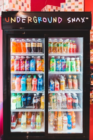 A display refrigerator filled with various bottled beverages, including sodas and juices, with a neon 'Underground Snax' sign on top. The bottles are organized neatly on several shelves, each containing different brands and colors.