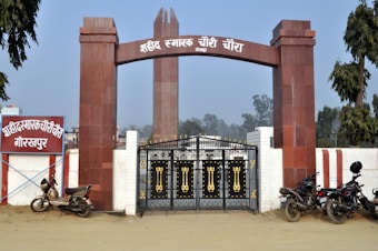 A large entrance with red and white columns and an archway, featuring a sign with text in an Indic script. Below the arch, there is a gate with ornate gold details. Several motorcycles are parked near the entrance, and trees are visible in the background.