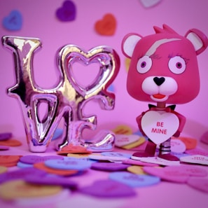 A pink bear figurine holding a heart with the words 'Be Mine' is surrounded by colorful heart-shaped candies. A metallic decoration spells out the word 'LOVE'. The background is also filled with heart shapes in various colors.