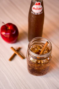 A clear glass jar filled with a brown, fizzy beverage is placed on a light wooden surface. Cinnamon sticks are visible in the drink and scattered on the table, adding to the rustic aesthetic. Nearby is a red apple and a bottle with a label reading 'Sidral Mundet', suggesting an apple-flavored soda theme.