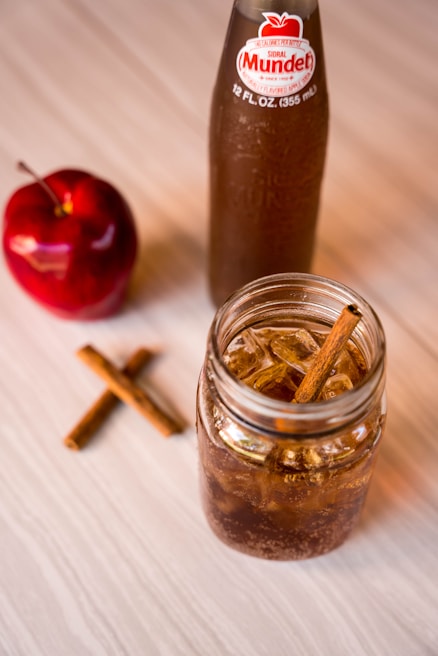 A clear glass jar filled with a brown, fizzy beverage is placed on a light wooden surface. Cinnamon sticks are visible in the drink and scattered on the table, adding to the rustic aesthetic. Nearby is a red apple and a bottle with a label reading 'Sidral Mundet', suggesting an apple-flavored soda theme.
